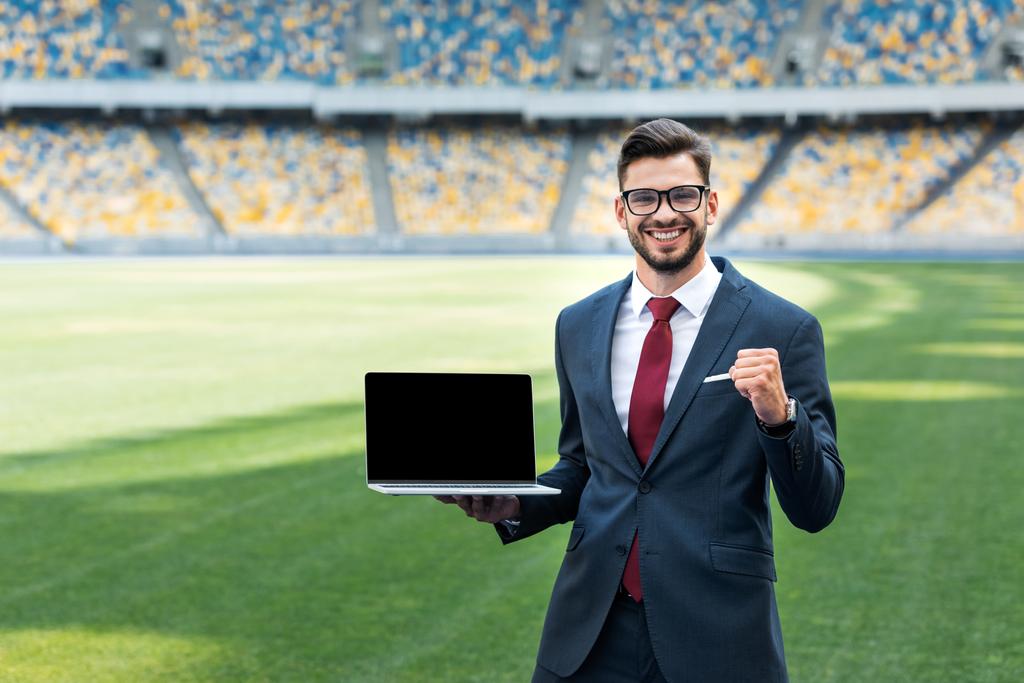 sonriente joven hombre de negocios en traje mostrando sí gesto mientras sostiene el ordenador portátil con pantalla en blanco en el estadio
 - Foto, Imagen
