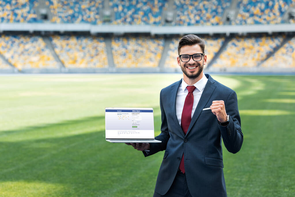 KIEW, UKRAINE - 20. JUNI 2019: Lächelnder junger Geschäftsmann im Anzug zeigt Ja-Geste, während er Laptop mit Facebook-Seite im Stadion hält - Foto, Bild