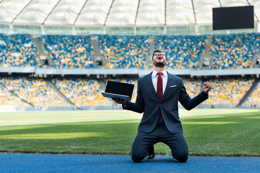 feliz joven hombre de negocios en traje mostrando sí gesto y la celebración de la computadora portátil con pantalla en blanco, mientras que de pie sobre las rodillas en el estadio
 - Foto, imagen