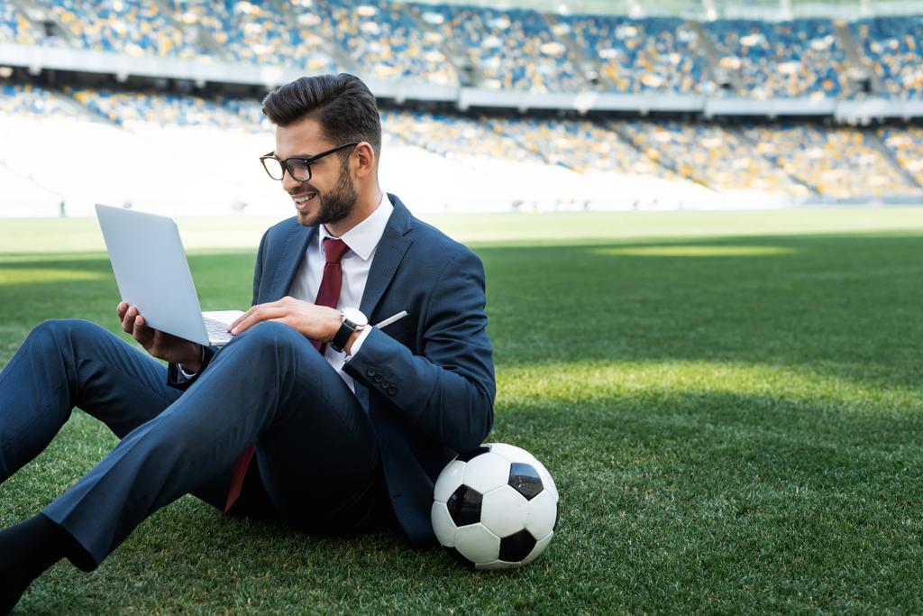 χαμογελαστός νεαρός επιχειρηματίας με κοστούμι με φορητό υπολογιστή και μπάλα ποδοσφαίρου κάθεται στο γήπεδο ποδοσφαίρου, έννοια αθλητικών στοιχημάτων - Φωτογραφία, εικόνα