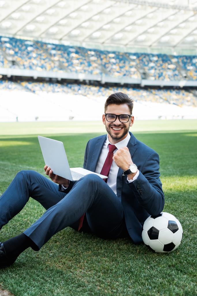 χαμογελαστός νεαρός επιχειρηματίας με κοστούμι με φορητό υπολογιστή και μπάλα ποδοσφαίρου κάθεται στο γήπεδο ποδοσφαίρου και δείχνει ναι χειρονομία στο γήπεδο, έννοια αθλητικών στοιχημάτων - Φωτογραφία, εικόνα