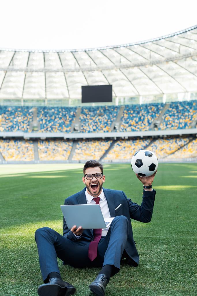 χαρούμενος νεαρός επιχειρηματίας με κοστούμι με φορητό υπολογιστή και μπάλα ποδοσφαίρου κάθεται στο γήπεδο ποδοσφαίρου, έννοια αθλητικών στοιχημάτων - Φωτογραφία, εικόνα