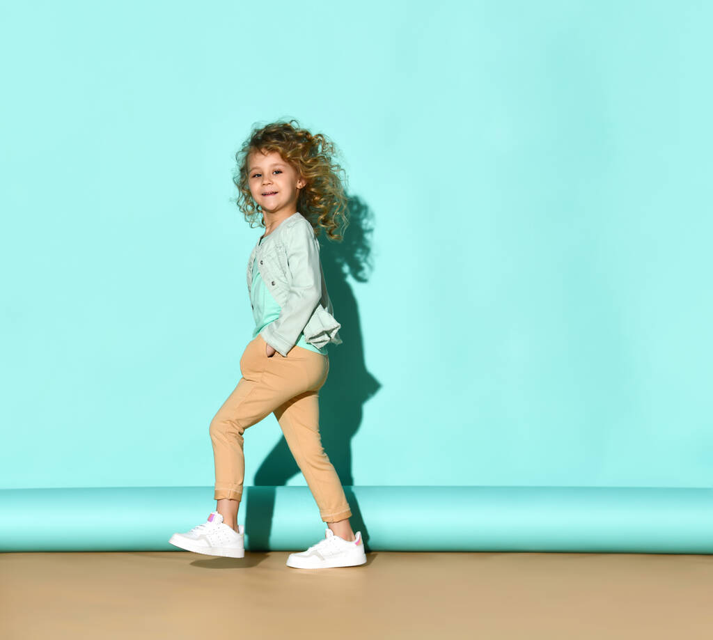 Petit enfant en jean, pantalon beige et baskets blanches. Elle sourit, les mains dans les poches, posant latéralement sur fond turquoise - Photo, image