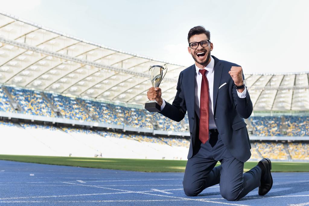 boldog fiatal üzletember öltönyben térden állva a futópályán trófeával a stadionban - Fotó, kép