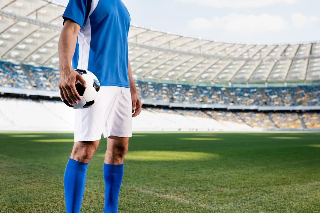 vista recortada de jugador de fútbol profesional en uniforme azul y blanco con pelota en el campo de fútbol en el estadio
 - Foto, imagen