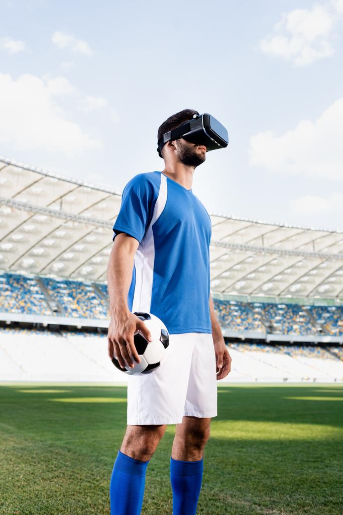 professionele voetballer in vr headset en blauw-wit uniform met bal op voetbalveld in het stadion - Foto, afbeelding