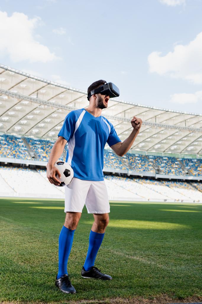 profesionální fotbalista ve vr headsetu a modré a bílé uniformě s míčkem křičí a ukazuje ano gesto na fotbalovém hřišti na stadionu - Fotografie, Obrázek
