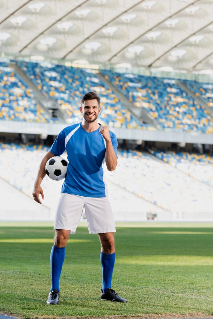 χαρούμενος επαγγελματίας ποδοσφαιριστής σε μπλε και άσπρη στολή με μπάλα δείχνει ναι χειρονομία στο γήπεδο ποδοσφαίρου - Φωτογραφία, εικόνα