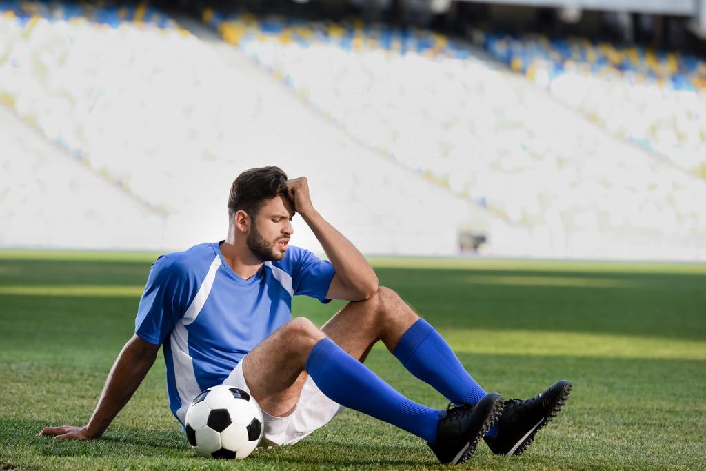 Trauriger Fußballprofi in blau-weißer Uniform sitzt mit Ball auf Fußballplatz im Stadion - Foto, Bild