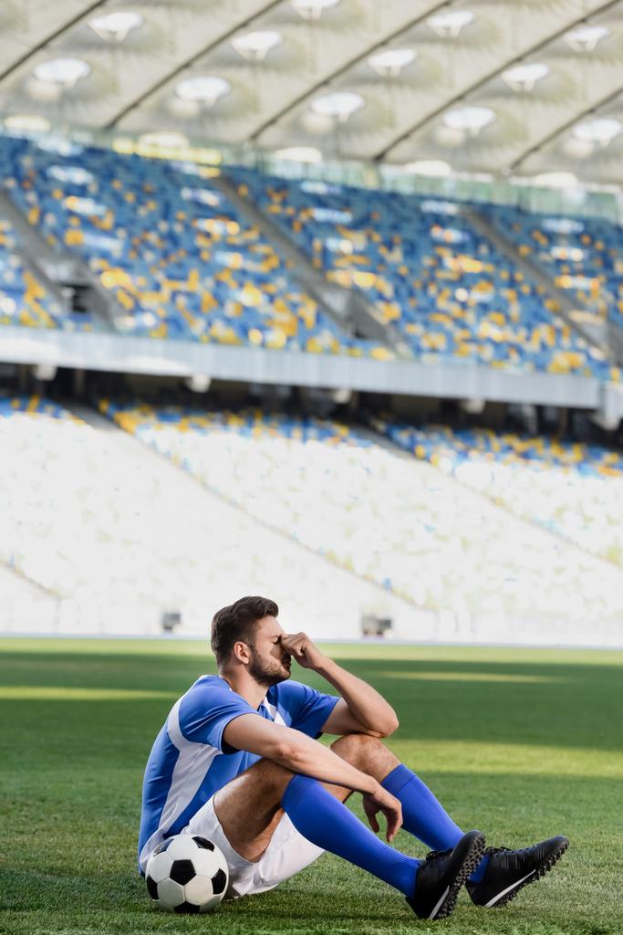 λυπημένος επαγγελματίας ποδοσφαιριστής σε μπλε και άσπρη στολή κάθεται με μπάλα στο γήπεδο ποδοσφαίρου - Φωτογραφία, εικόνα