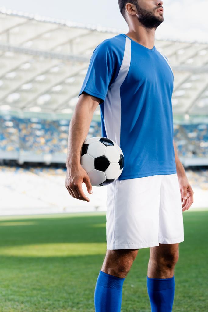 oříznutý pohled profesionálního fotbalisty v modré a bílé uniformě s míčem na fotbalovém hřišti na stadionu - Fotografie, Obrázek