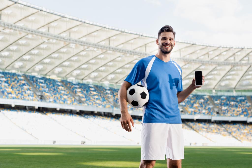 улыбающийся профессиональный футболист в сине-белой форме с мячом, показывающим смартфон с пустым экраном на стадионе
 - Фото, изображение
