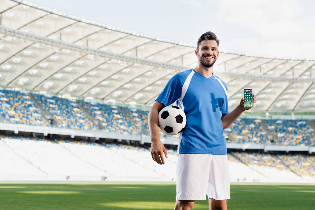 KYIV, UKRAINE - czerwiec 20, 2019: uśmiechnięty profesjonalny piłkarz w niebiesko-białym mundurze z piłką pokazującą smartfona z aplikacjami na iPhone 'a na stadionie - Zdjęcie, obraz