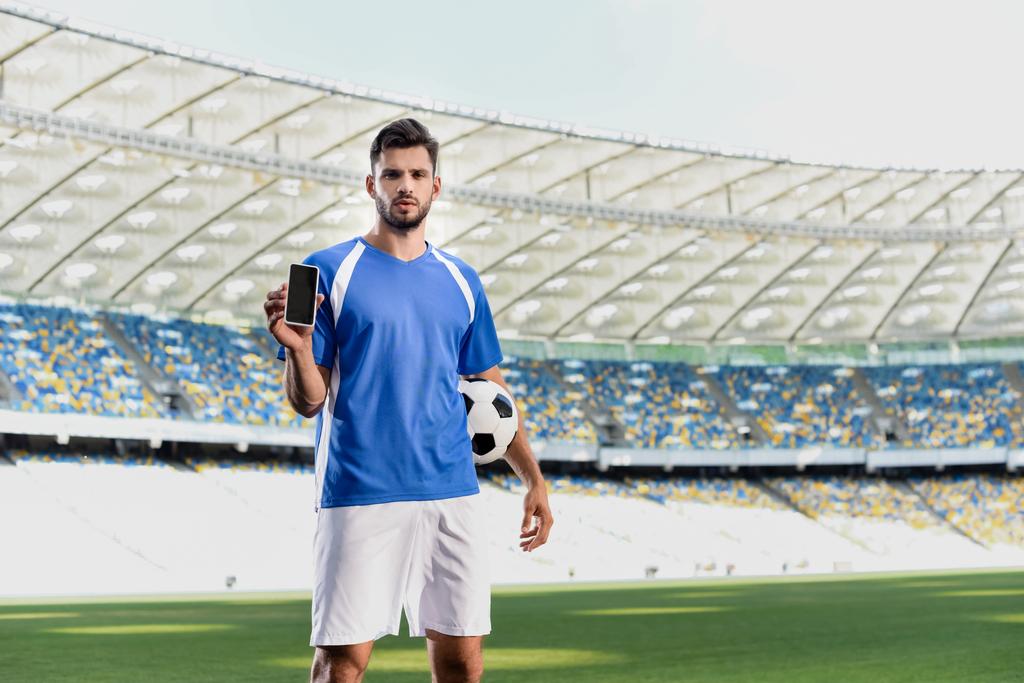professionele voetballer in blauw en wit uniform met bal tonen smartphone met leeg scherm in het stadion - Foto, afbeelding