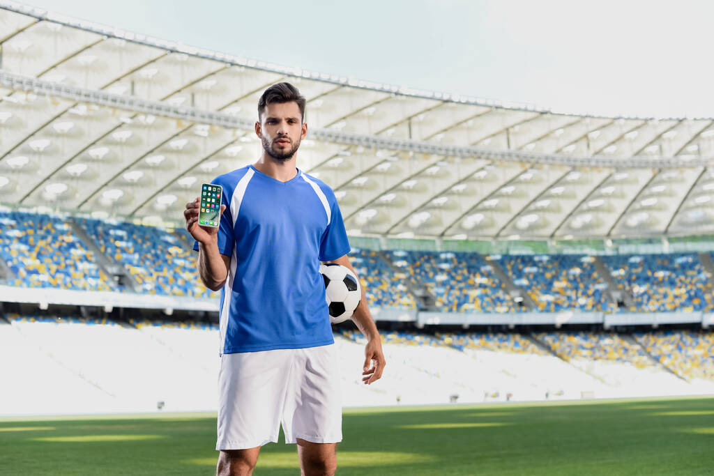 KYIV, UCRANIA - 20 de junio de 2019: futbolista profesional en uniforme azul y blanco con pelota que muestra el teléfono inteligente con aplicaciones de iphone en el estadio
 - Foto, imagen