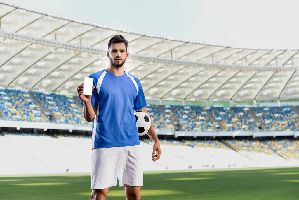 επαγγελματίας ποδοσφαιριστής σε μπλε και άσπρη στολή με μπάλα δείχνει smartphone με λευκή οθόνη στο γήπεδο - Φωτογραφία, εικόνα