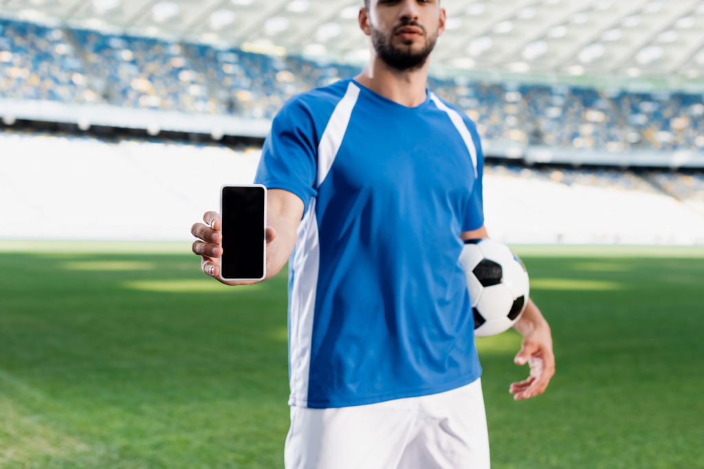 cropped view of professional soccer player in blue and white uniform with ball showing smartphone with blank screen at stadium - Photo, Image