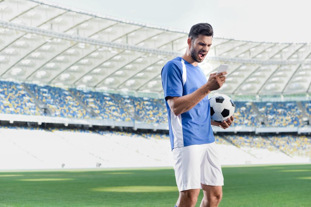 emotionele professionele voetballer in blauw en wit uniform met bal met smartphone in het stadion - Foto, afbeelding
