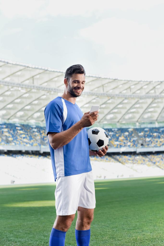 uśmiechnięty profesjonalny piłkarz w niebiesko-białym mundurze z piłką przy użyciu smartfona na stadionie - Zdjęcie, obraz