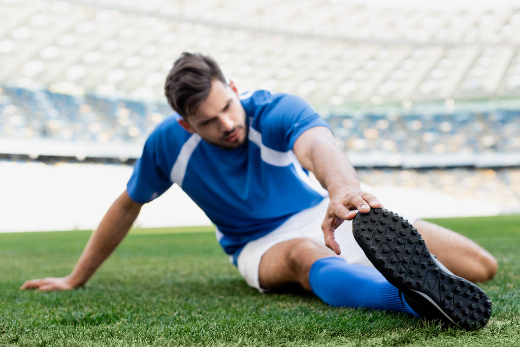 selective focus of professional soccer player in blue and white uniform stretching on football pitch at stadium - Photo, Image