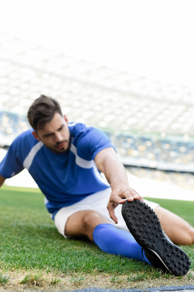 επιλεκτική εστίαση του επαγγελματία ποδοσφαιριστή σε μπλε και άσπρη στολή που εκτείνεται στο γήπεδο ποδοσφαίρου στο γήπεδο - Φωτογραφία, εικόνα