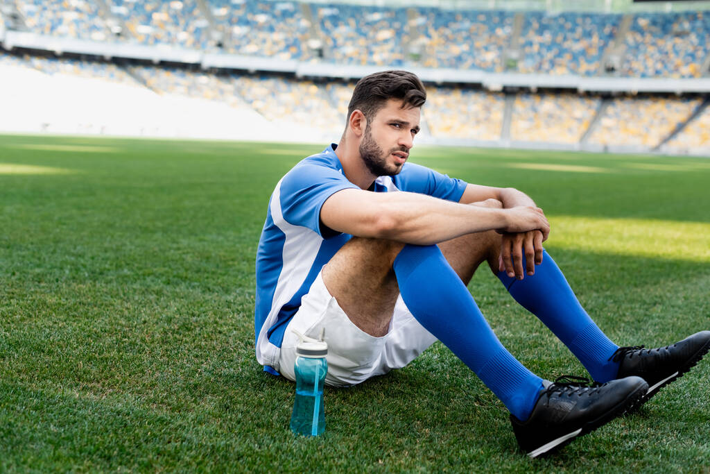 プロのサッカー選手は青と白のユニフォームを着てスタジアムのスポーツボトルとサッカーピッチに座って ロイヤリティフリー写真 画像素材