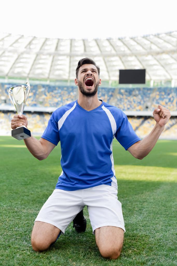 Fußballprofi in blau-weißer Uniform mit Sportpokal steht auf Knien auf Fußballplatz und brüllt im Stadion - Foto, Bild