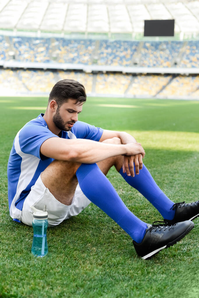 zawodowy piłkarz w niebiesko-białym mundurze siedzący na boisku do piłki nożnej z butelką sportową na stadionie - Zdjęcie, obraz