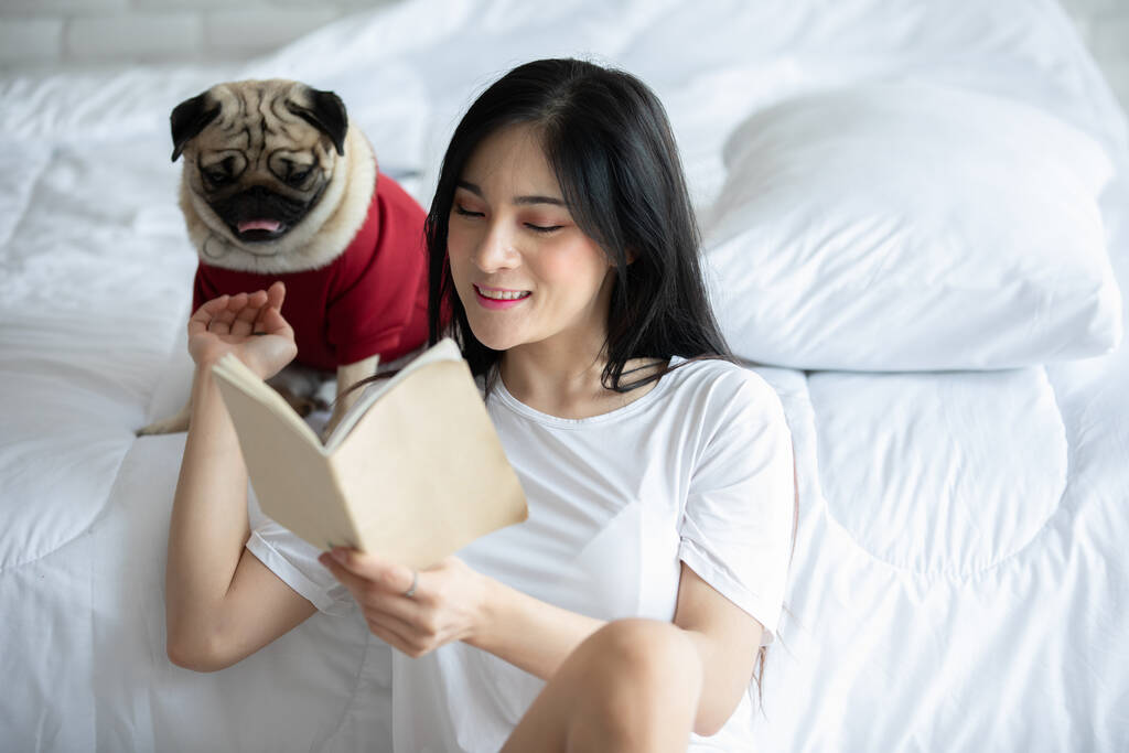 Houkutteleva aasialainen nuori nainen lukee kirjaa, kun hänen koiransa mopsi rotu viihtyisässä makuuhuoneessa rentouttava ja mukava lomilla, Virkistys koiran kanssa Concept
 - Valokuva, kuva