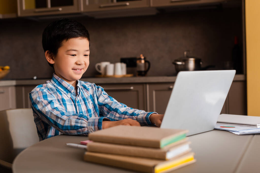 hymyilevä aasialainen poika opiskelee verkossa kannettavan tietokoneen ja kirjojen kanssa kotona karanteenin aikana - Valokuva, kuva