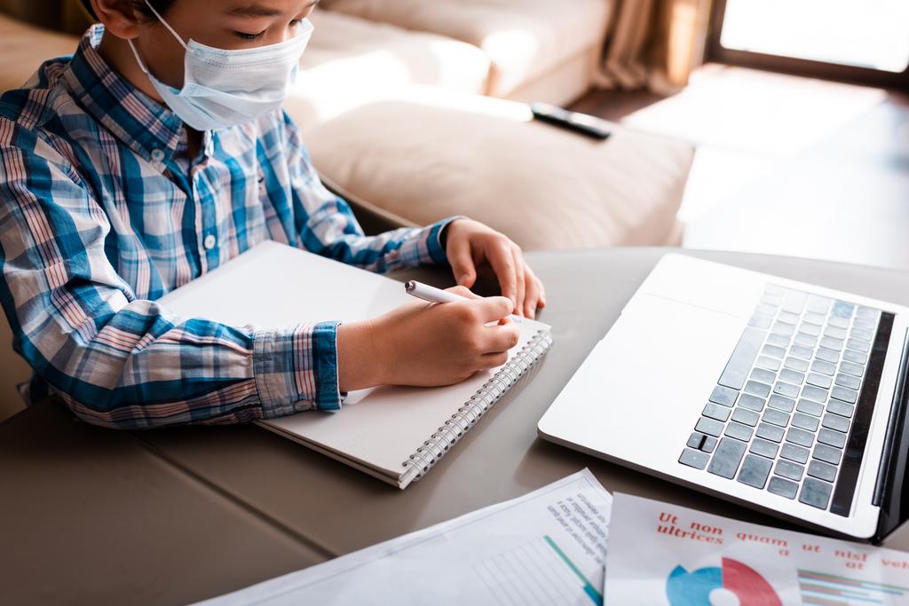 Мальчик в медицинской маске пишет и учится онлайн с ноутбуком дома во время самоизоляции - Фото, изображение