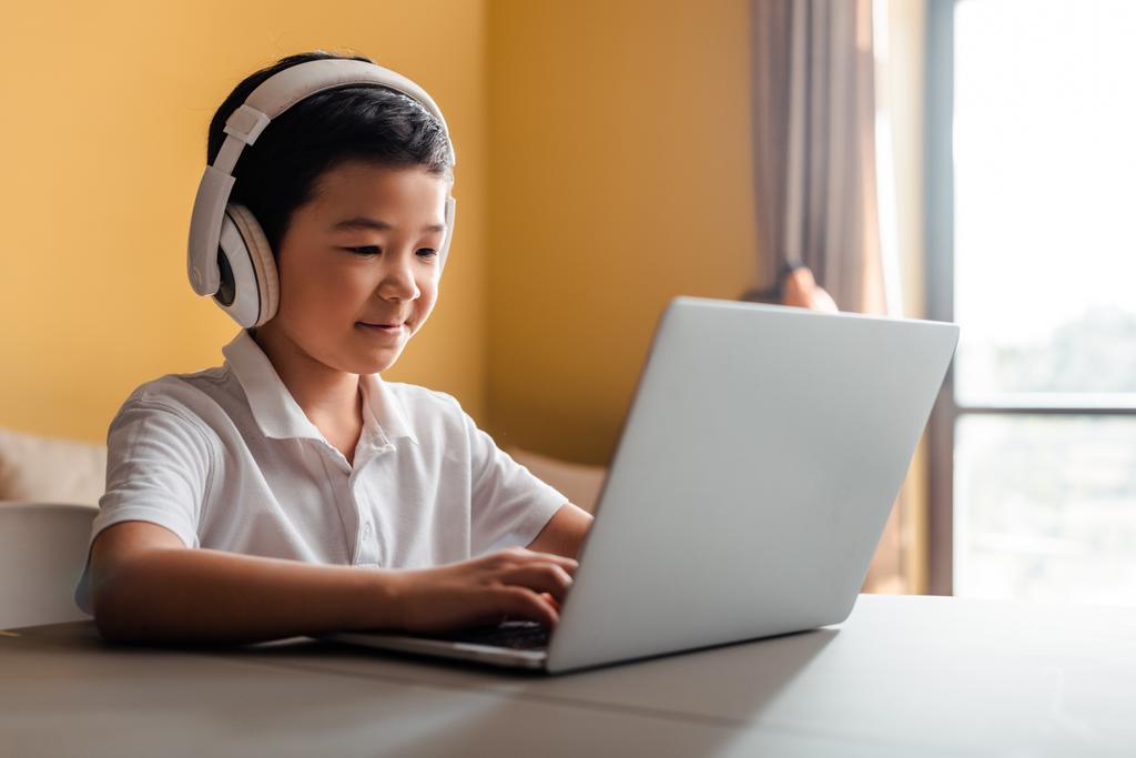 lächelnder asiatischer Junge beim Online-Lernen mit Laptop und Kopfhörer zu Hause während der Quarantäne - Foto, Bild