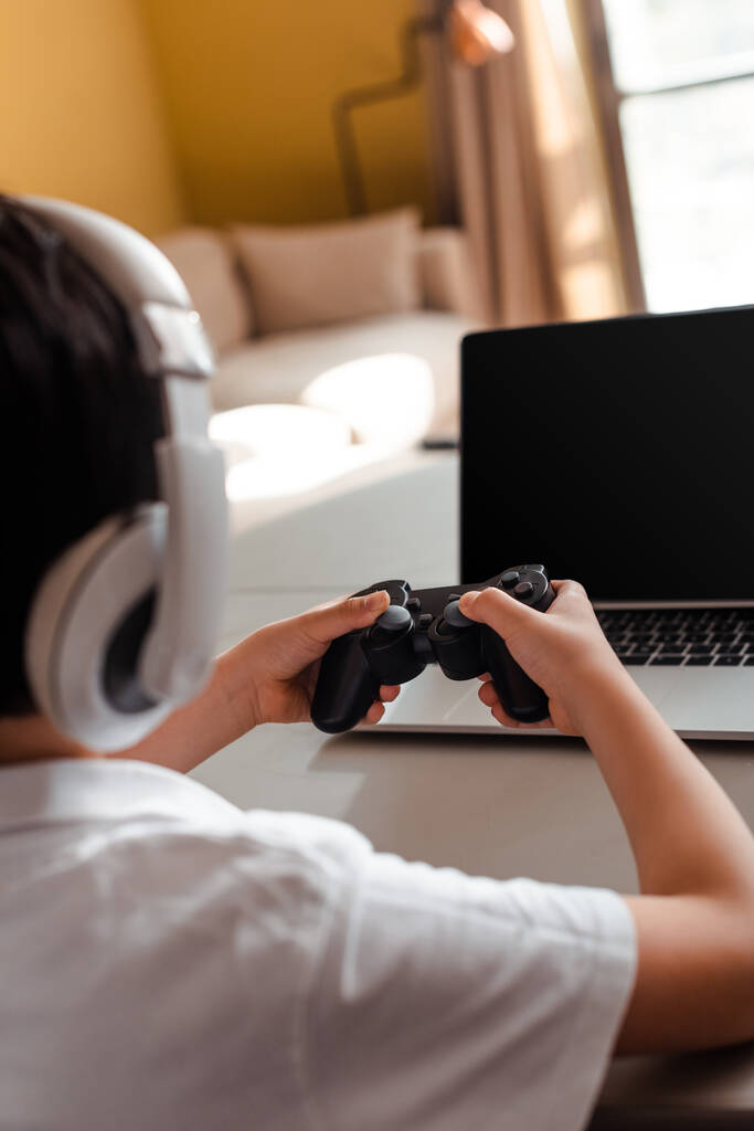 KYIV, UKRAINE - APRIL 22, 2020: cropped view on little boy playing video game with joystick and laptop with blank screen on quarantine - Photo, Image