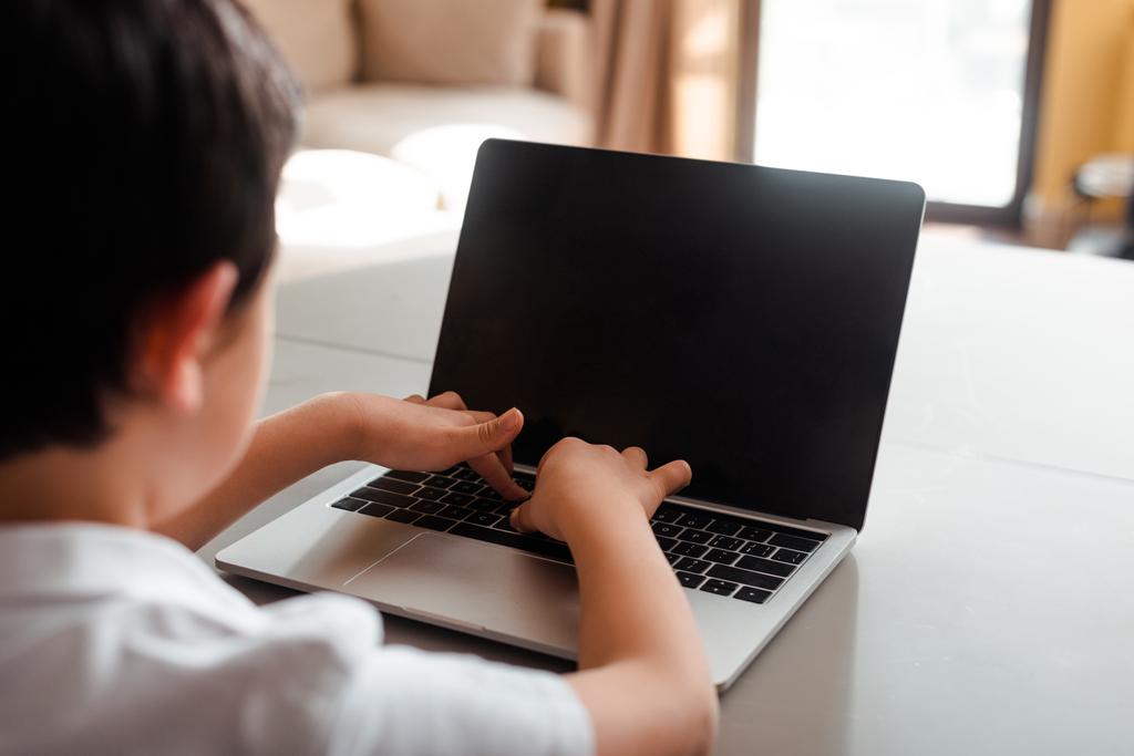 καλλιεργημένη άποψη του αγοριού που σπουδάζει online και δακτυλογράφηση σε φορητό υπολογιστή με κενή οθόνη στο σπίτι κατά τη διάρκεια της αυτο-απομόνωσης - Φωτογραφία, εικόνα