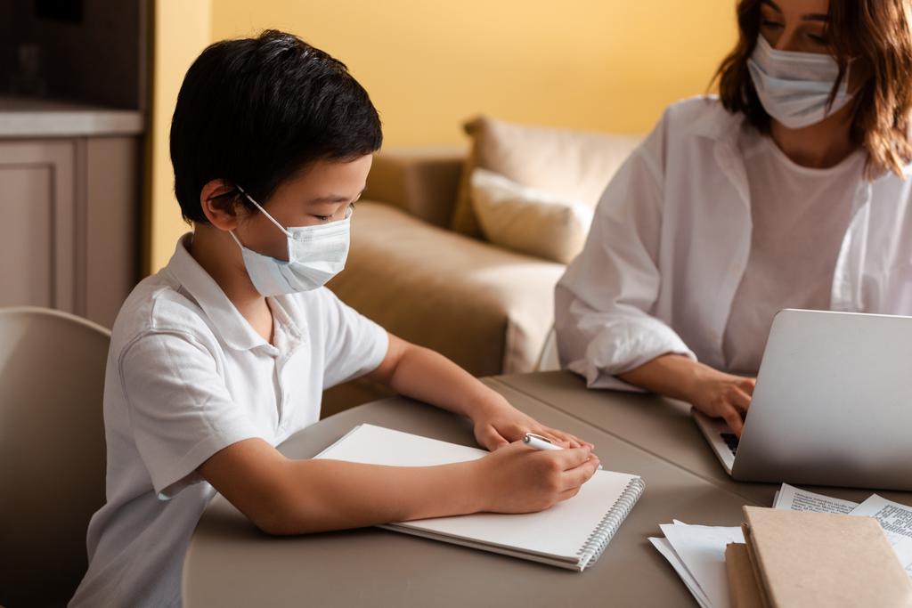 schattig aziatisch jongen in medisch masker studeren met moeder thuis op quarantaine - Foto, afbeelding