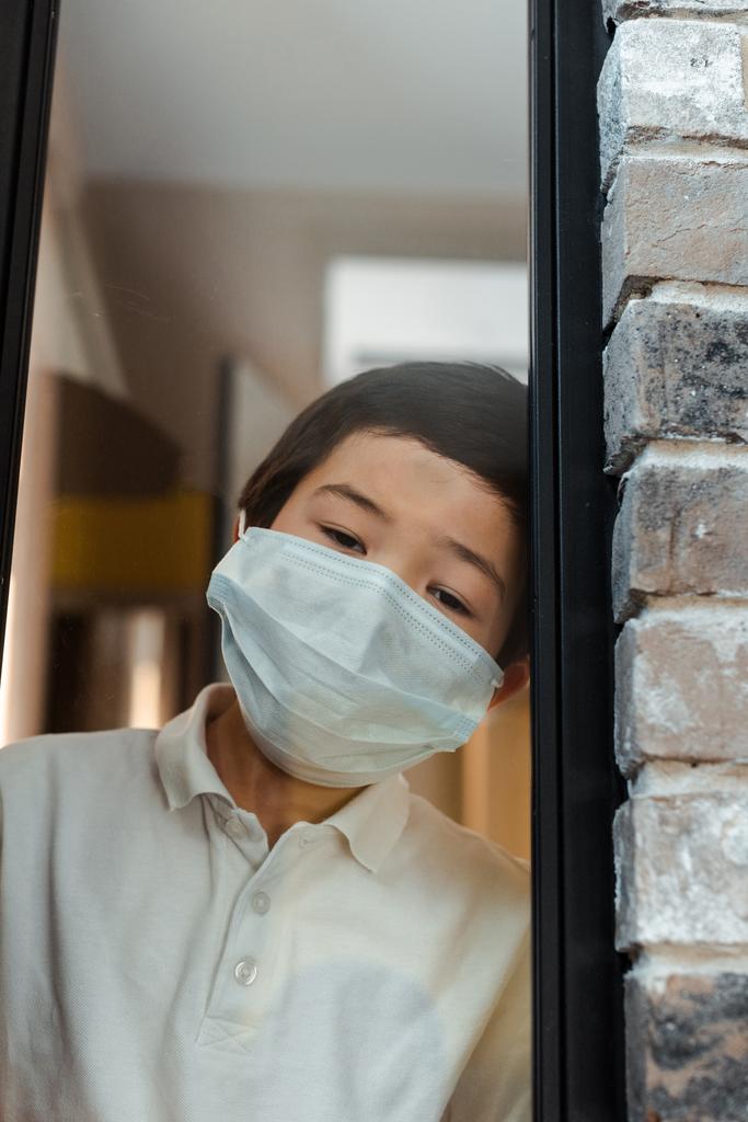 αναστατωμένος ασιατικό αγόρι σε ιατρική μάσκα κοιτάζοντας μέσα από το παράθυρο στην καραντίνα - Φωτογραφία, εικόνα