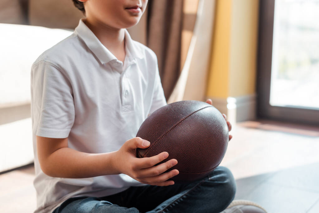 vista panorámica del niño asiático con balón de rugby sentado en el suelo en casa en cuarentena.  - Foto, Imagen