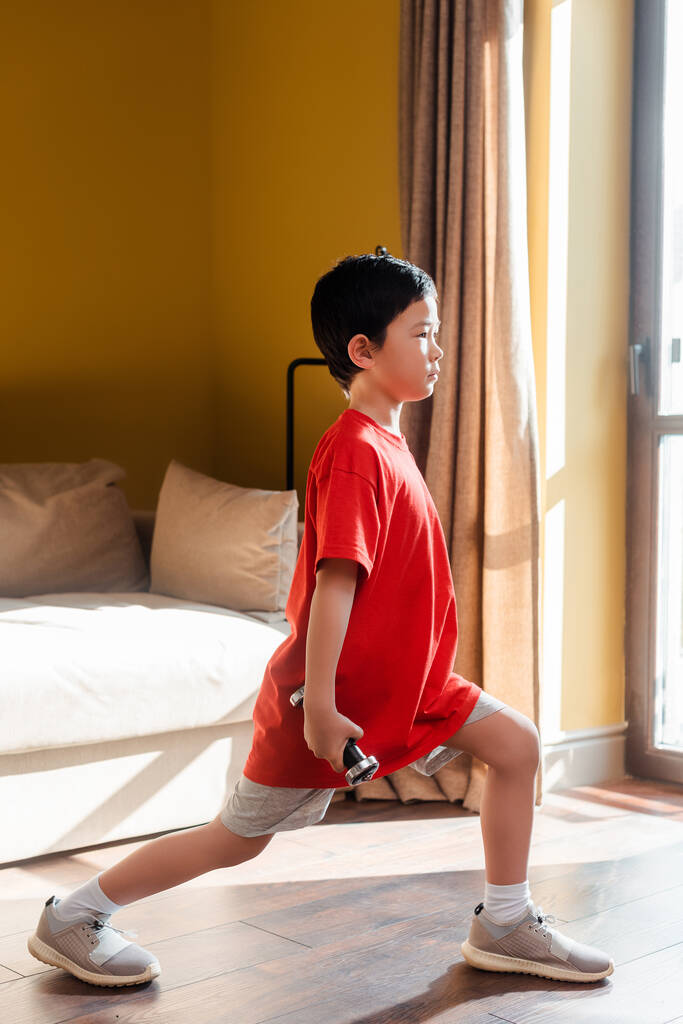 sportlicher asiatischer Junge macht während der Quarantäne zu Hause Ausfallversuche mit Kurzhanteln - Foto, Bild