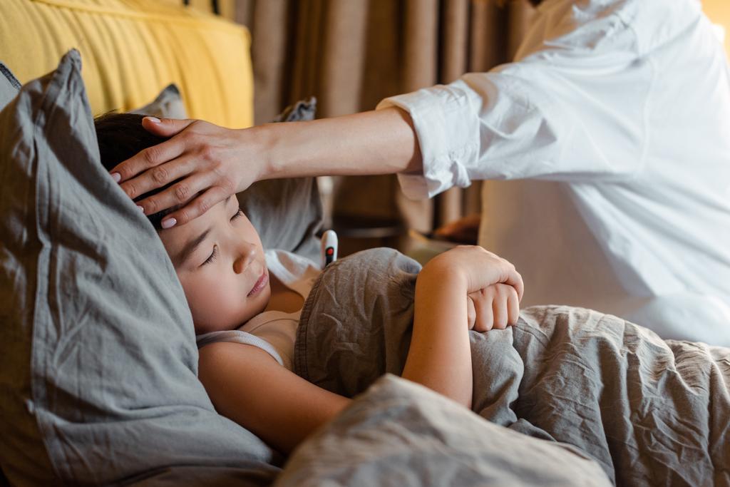 beteg ázsiai fiú a hőmérséklet feküdt az ágyban anya közel karantén - Fotó, kép