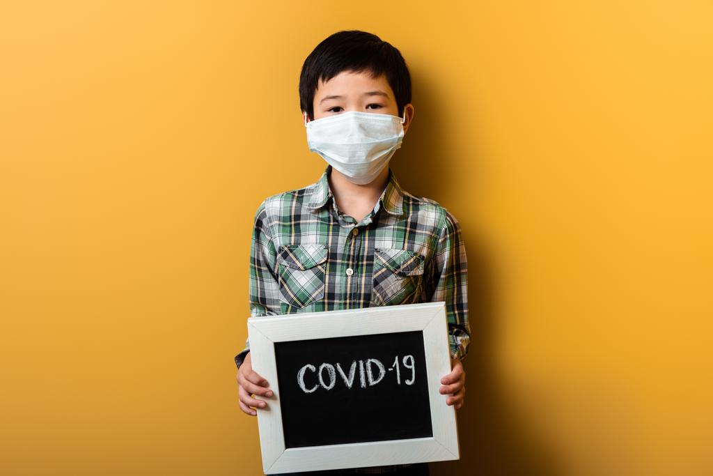 azjatycki chłopiec w masce medycznej trzymając pokładzie z covid-19 znak na żółty podczas kwarantanny - Zdjęcie, obraz