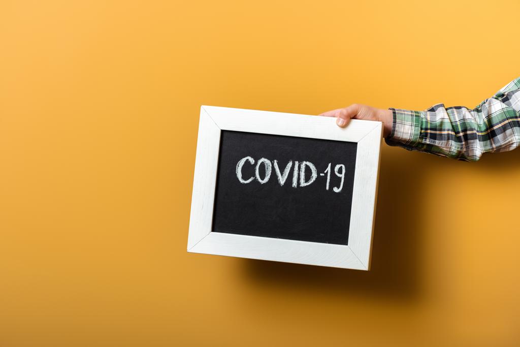 bijgesneden weergave van jongen holding board met covid-19 teken op geel tijdens zelfisolatie - Foto, afbeelding