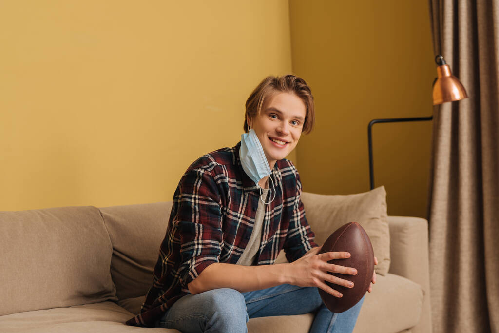 felice giovane uomo in maschera medica che tiene il calcio americano in soggiorno
 - Foto, immagini