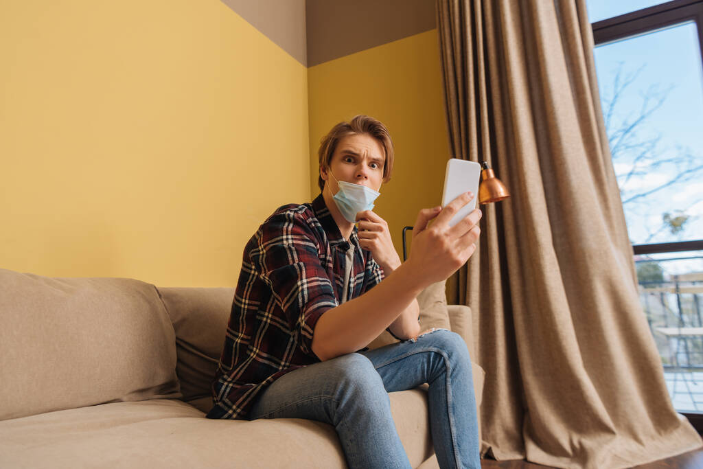 σοκαρισμένος άνθρωπος με ιατρική μάσκα κοιτάζοντας smartphone στο σαλόνι  - Φωτογραφία, εικόνα
