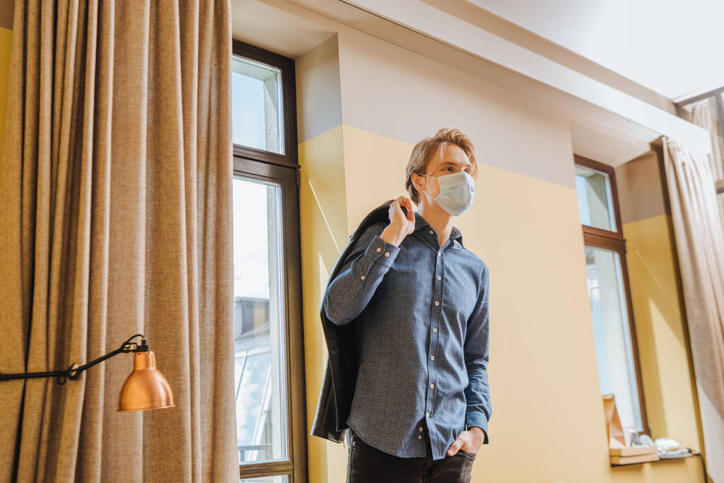muž v lékařské masce drží bundu a stojí s rukou v kapse doma, konec koncepce karantény  - Fotografie, Obrázek