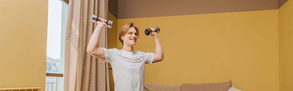πανοραμική καλλιέργεια ευτυχισμένη νεαρός άνδρας άσκηση με αλτήρες στο σαλόνι, τέλος της έννοιας καραντίνα - Φωτογραφία, εικόνα