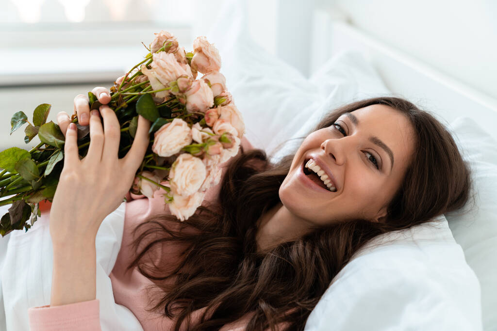 Εικόνα ενός χαρούμενου θετική όμορφη νεαρή γυναίκα σε εσωτερικούς χώρους στο σπίτι βρίσκεται στο υπνοδωμάτιο στο κρεβάτι κρατώντας λουλούδια. - Φωτογραφία, εικόνα