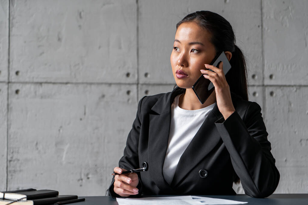 Serious asiática mujer de negocios en teléfono tomando notas
 - Foto, Imagen