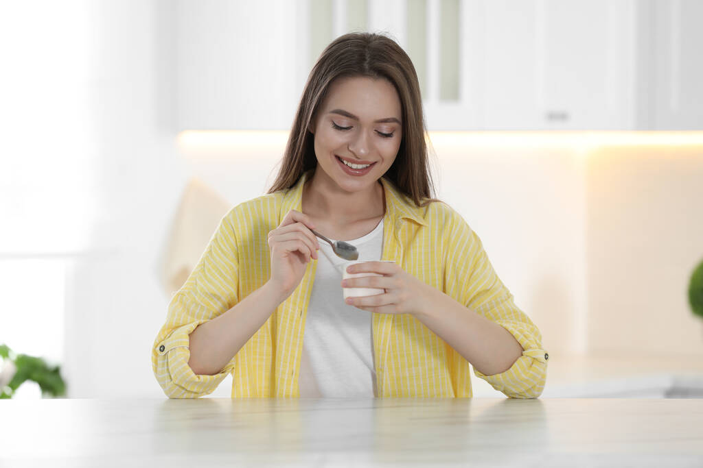 Молодая привлекательная женщина ест вкусный йогурт за столом на кухне
 - Фото, изображение