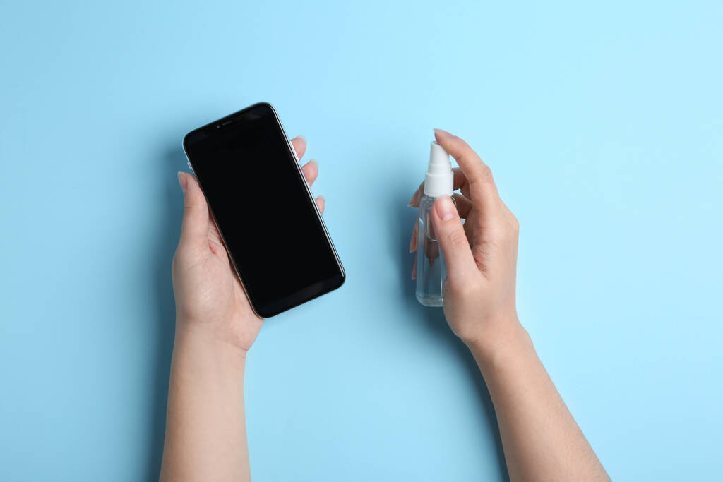 Женщина распыляет антисептик на смартфон на голубом фоне, вид сверху
 - Фото, изображение