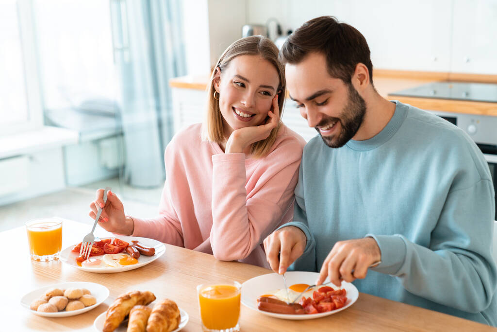 Porträt eines jungen glücklichen Paares, das gemeinsam am Tisch isst und zu Hause in der gemütlichen Küche frühstückt - Foto, Bild
